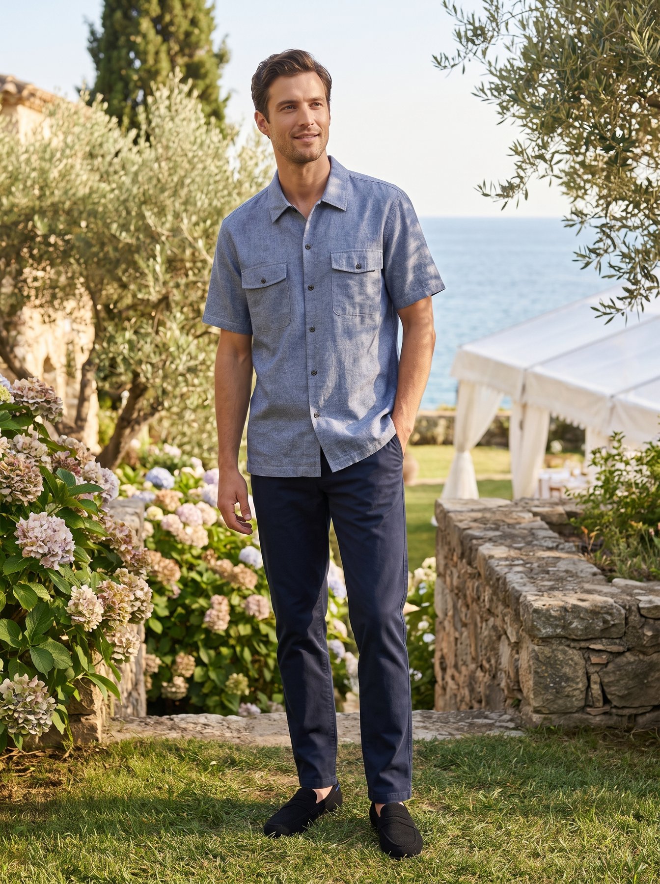 A man in a blue short-sleeve chambray shirt, tailored navy trousers and black suede penny loafers at a coastal summer wedding